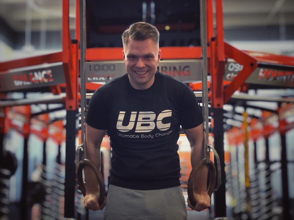 Uśmiechnięty mężczyzna w czarnej koszulce UBC trzyma się kółek gimnastycznych na tle pomarańczowej konstrukcji w siłowni.