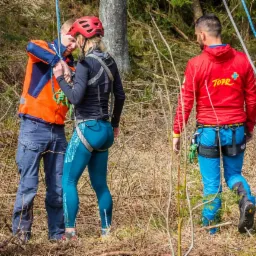Przygotowania do wspinaczki: Instruktor asekuruje uczestniczkę w czerwonym kasku, obok stoi mężczyzna w czerwonej kurtce z logo TOPR na tle leśnym.