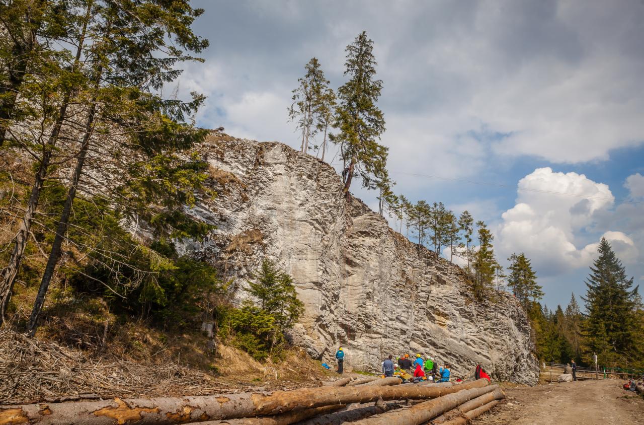 Grupa osób w kolorowych ubraniach odpoczywa u podnóża skalnej ściany z rosnącymi na niej drzewami, w tle widoczne niebo z chmurami.