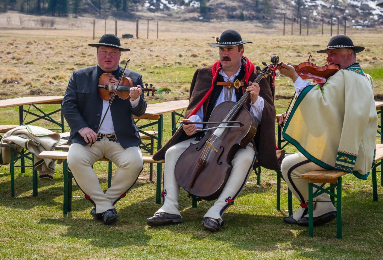 Trzech mężczyzn w strojach regionalnych grających na skrzypcach i wiolonczeli siedząc na ławce na tle łąki.