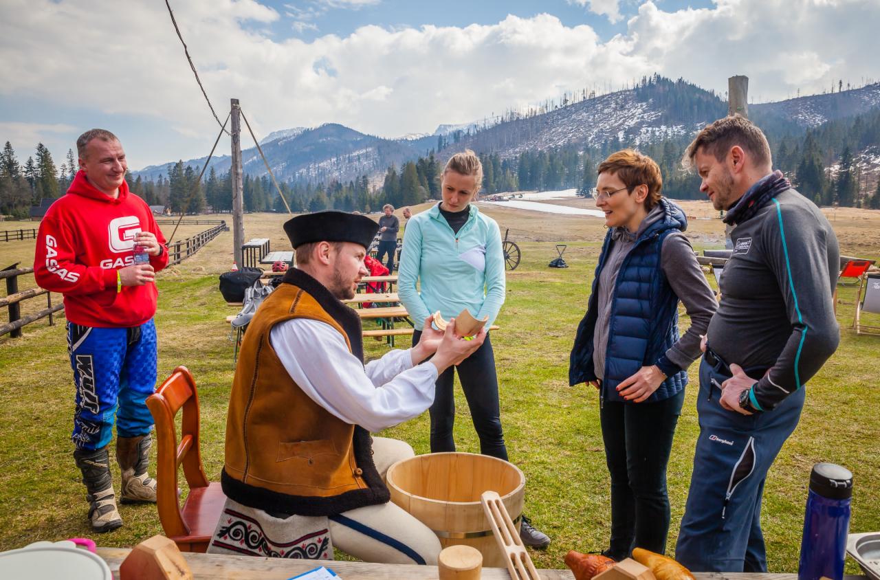 Mężczyzna w stroju regionalnym prezentuje drewniane naczynie grupie osób na tle górskiego krajobrazu, prawdopodobnie podczas pokazu lub warsztatów rzemiosła.