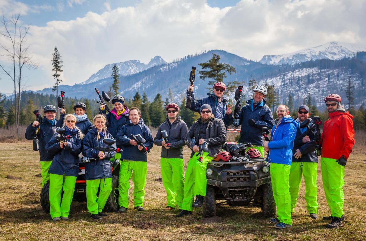Grupa osób w neonowych spodniach i z markerami paintballowymi pozuje na tle gór i quada.