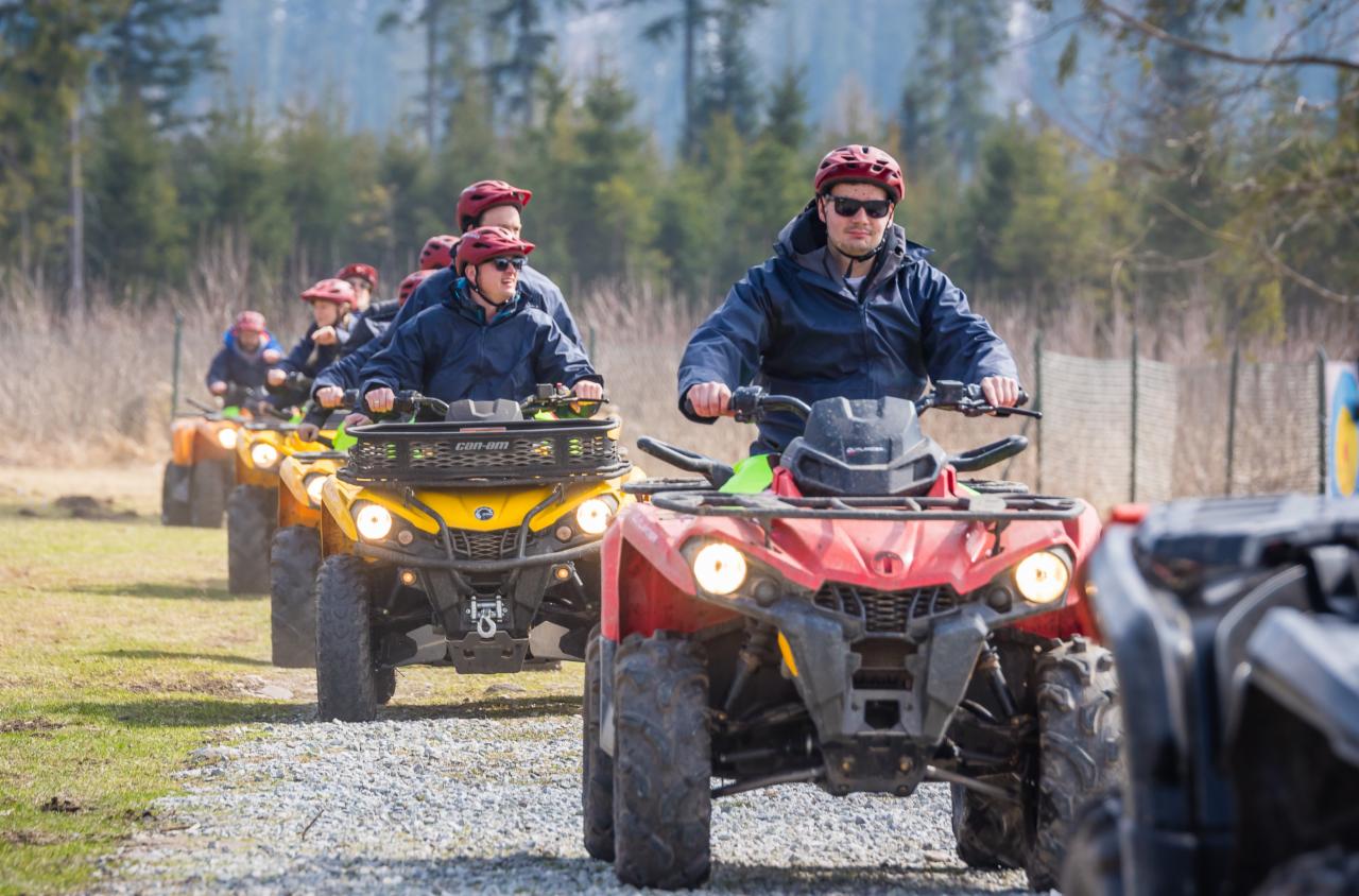Grupa osób w kaskach i okularach przeciwsłonecznych jedzie na quadach po szutrowej drodze w kierunku widza, w tle las iglasty i pagórki.