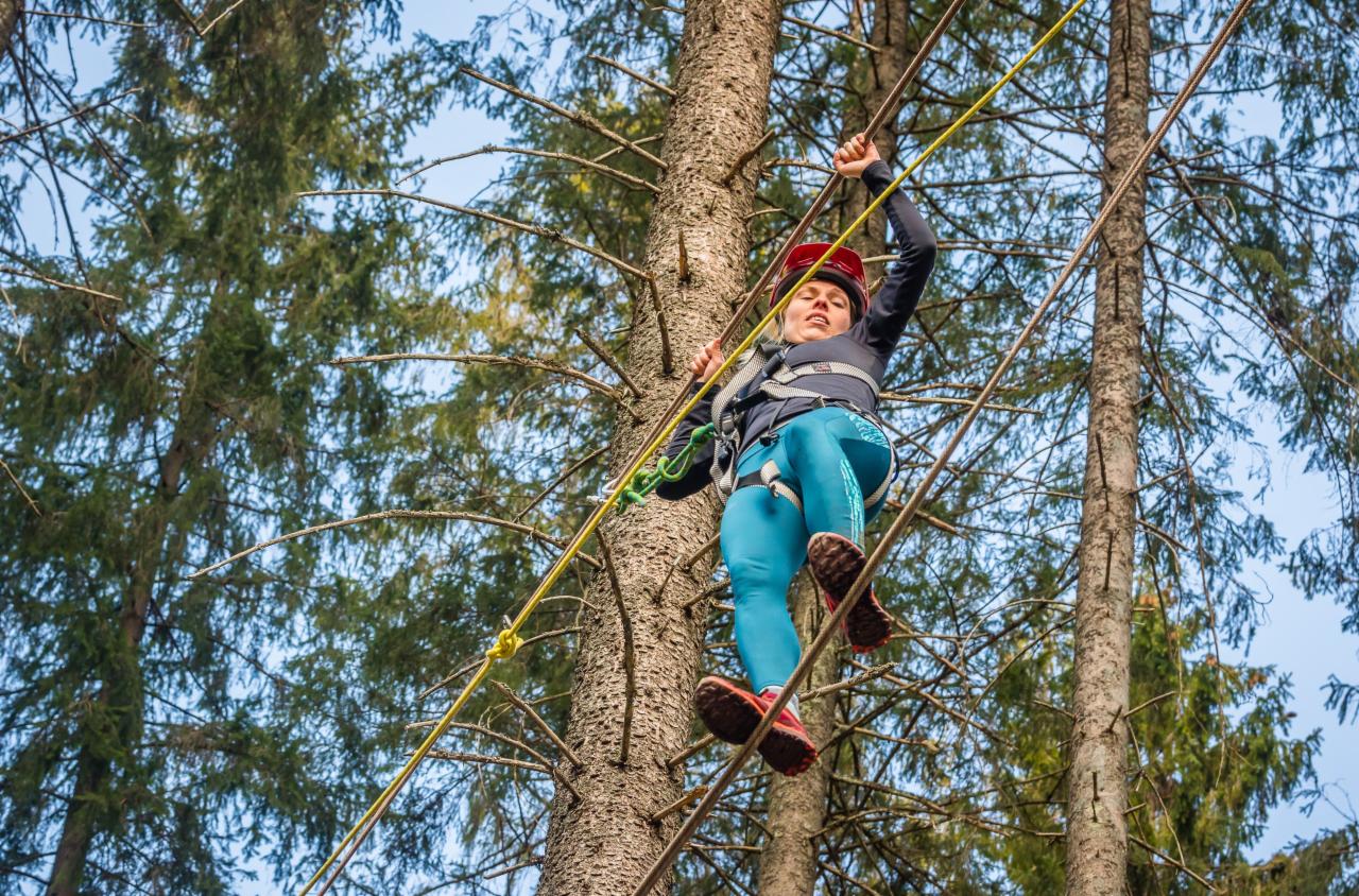 Uczestniczka w czerwonym kasku pokonuje przeszkodę linową w parku przygody, w tle wysokie drzewa iglaste i niebo.