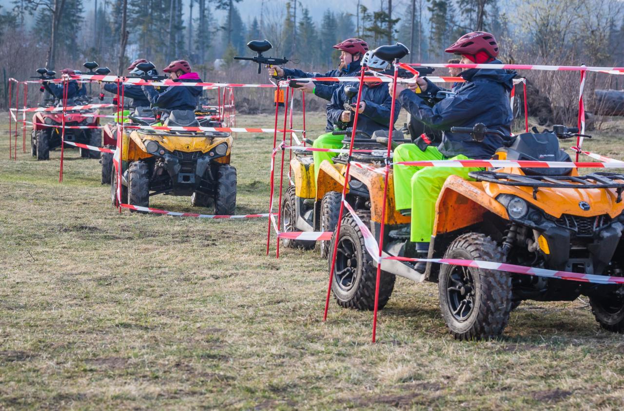 Grupa osób w kaskach i jaskrawych spodniach, siedzących na quadach i celujących z pistoletów do paintballa, oddzielona taśmą ostrzegawczą na tle leśnym.