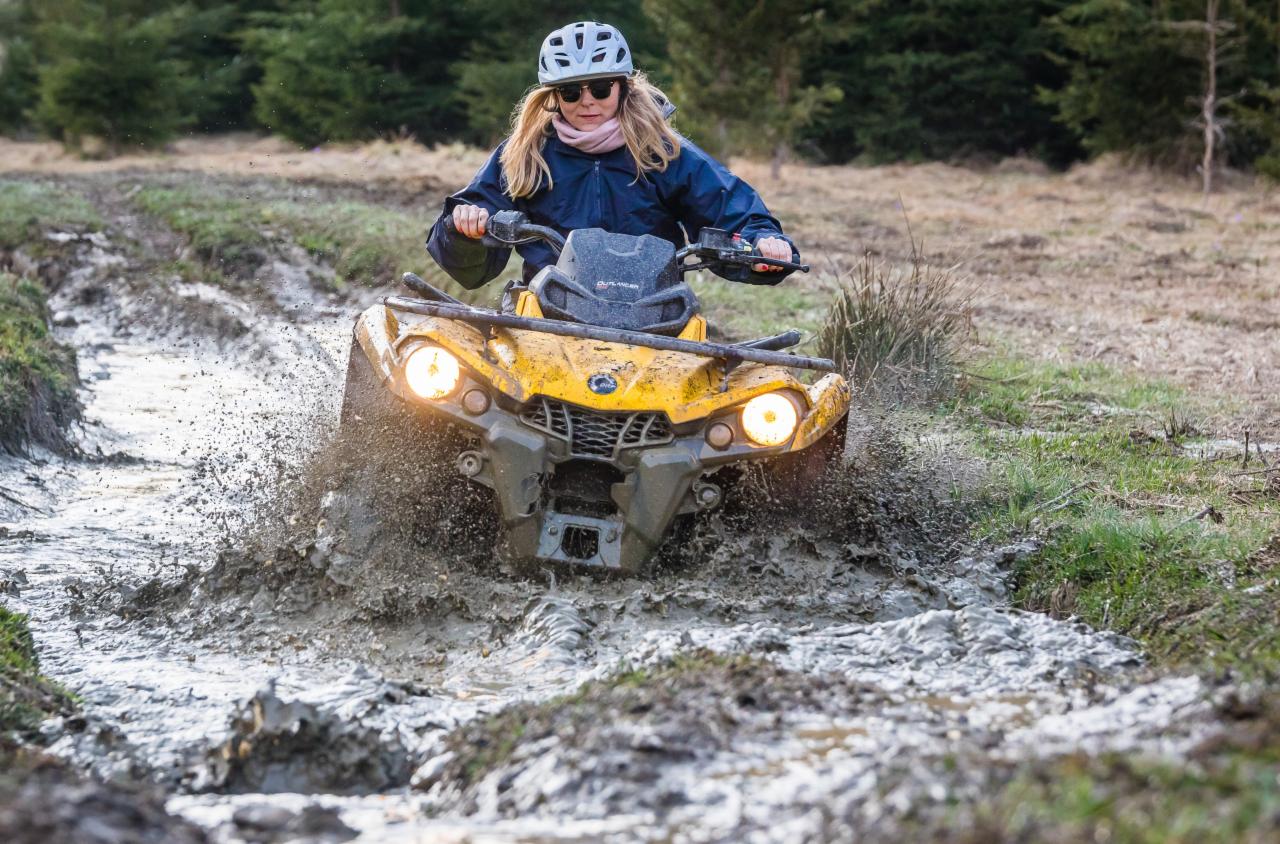 Kobieta w kasku i okularach przeciwsłonecznych prowadzi żółty quad przez błotnisty teren, z widocznymi rozpryskami błota i lasem w tle.