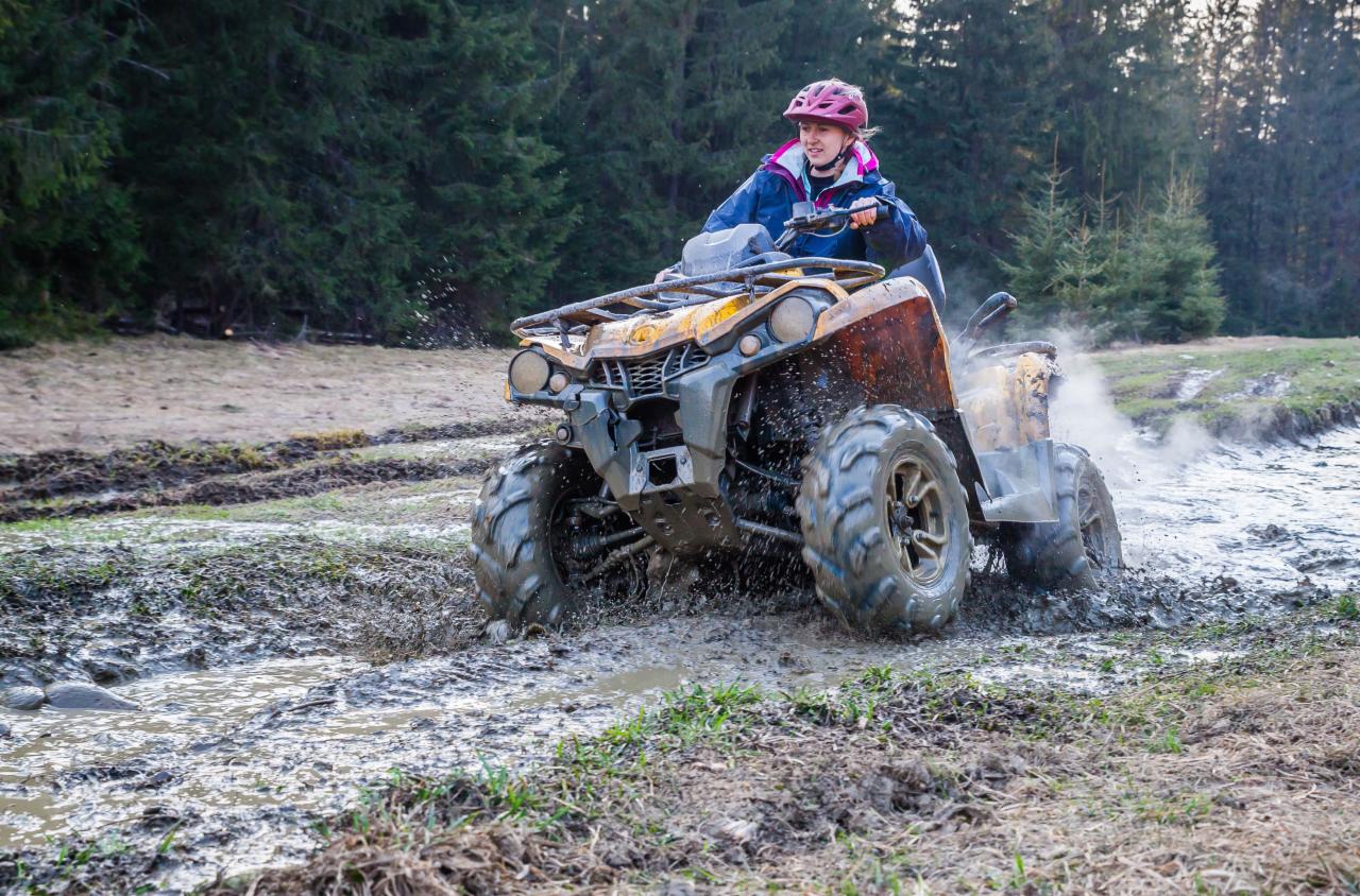 Kobieta w kasku na pomarańczowym quadzie przedziera się przez błotnistą kałużę, w tle las iglasty.