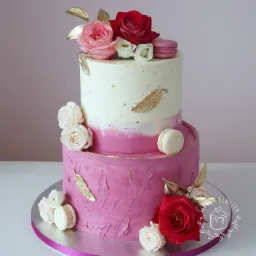 Dwupiętrowy tort zdobiony różowymi i białymi różami, makaronikami oraz złotymi piórkami, na srebrnej podkładce z różową wstążką.