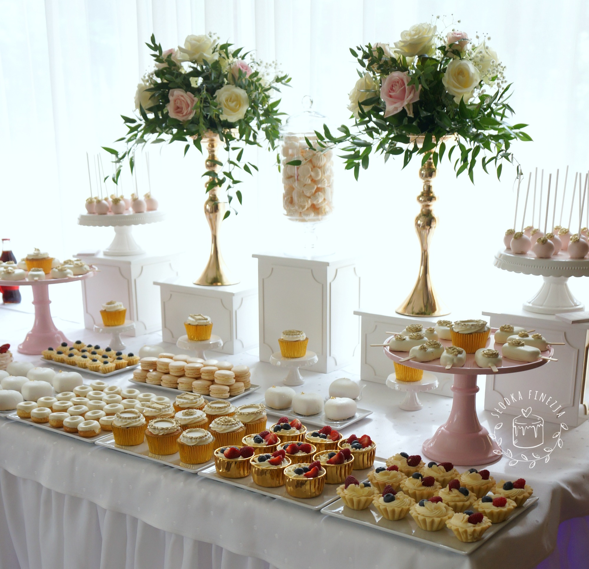 Elegancki stół z deserami na przyjęciu: babeczki z owocami, makaroniki, cake pops, mini donuty, białe desery, wszystko udekorowane na białym obrusie z kwiatami w złotych wazonach.