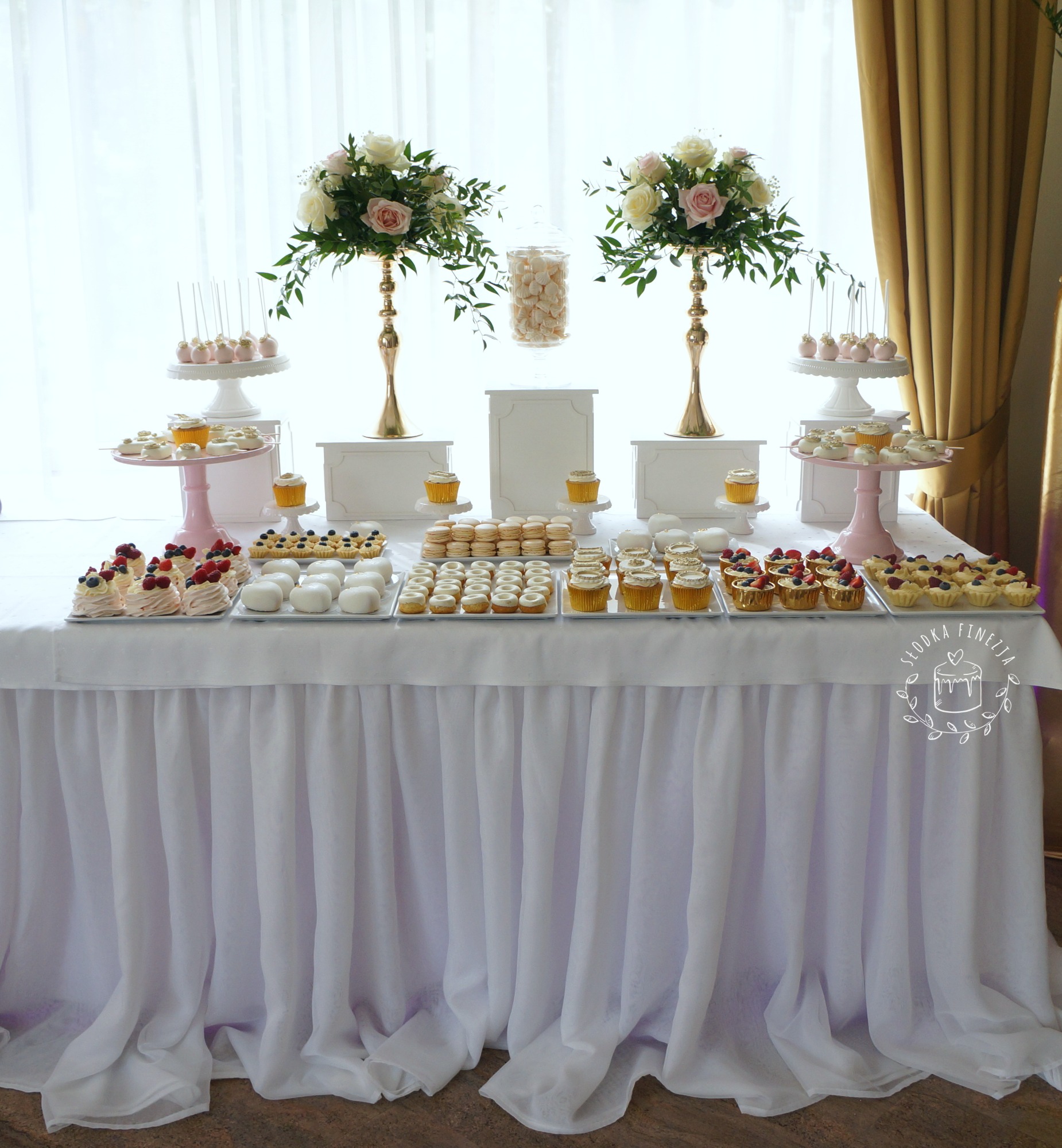 Stół z białym obrusem udekorowany kompozycjami kwiatowymi w złotych wazonach i różnorodnymi deserami: babeczki, cake pops, bezy z owocami, makaroniki.