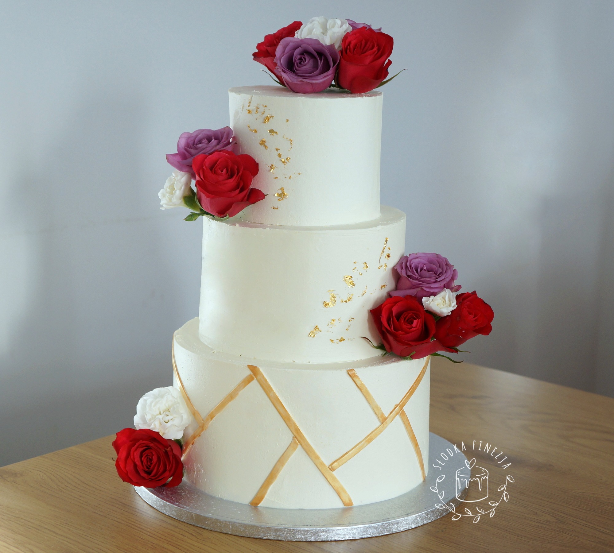 Trzypiętrowy tort weselny z białym kremem, zdobiony złotymi akcentami i kompozycją z czerwonych, białych i fioletowych róż, umieszczony na srebrnej podkładce na drewnianym stole, z widocznym logo...
