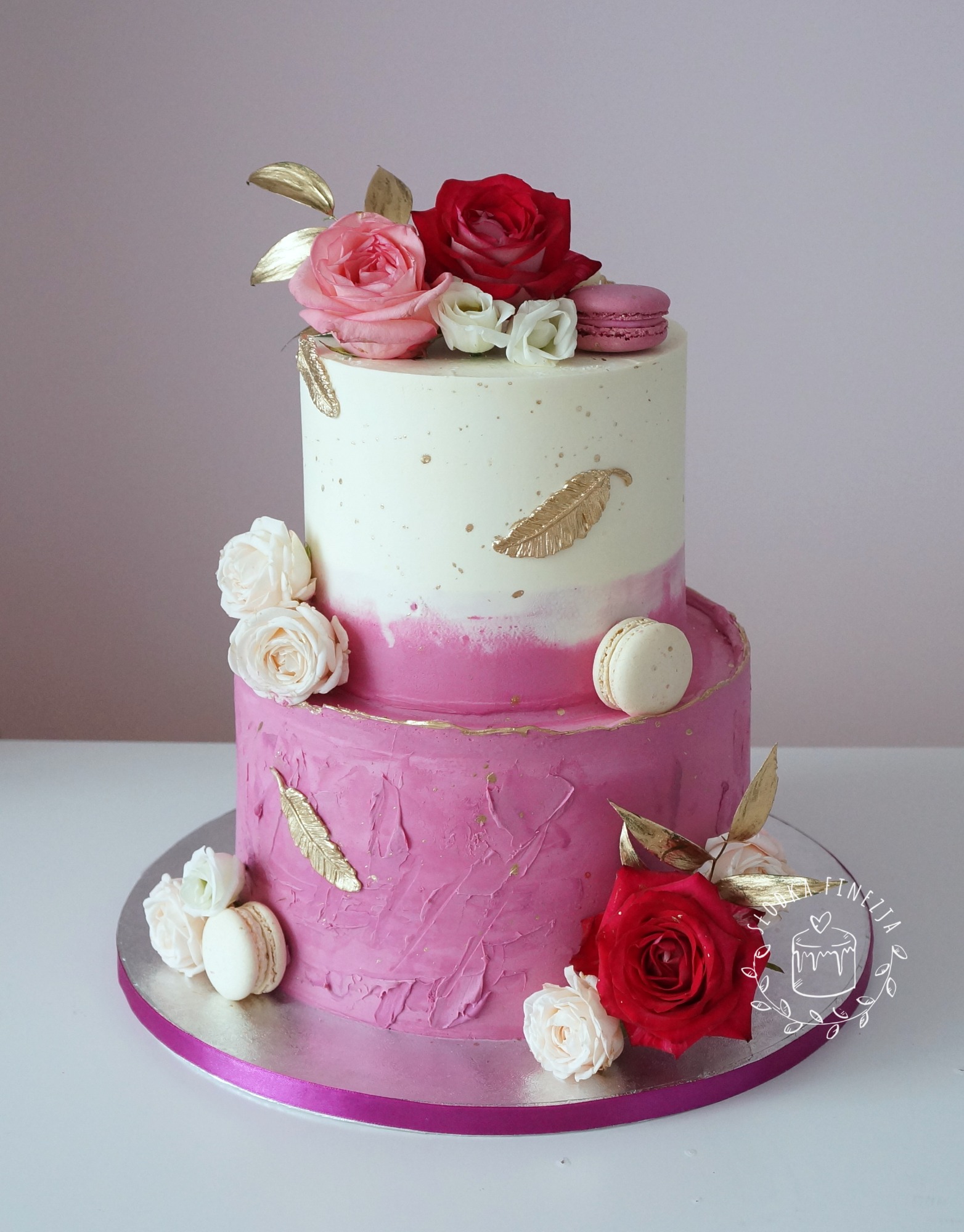 Dwupiętrowy tort zdobiony różowymi i białymi różami, makaronikami oraz złotymi piórkami, na srebrnej podkładce z różową wstążką.