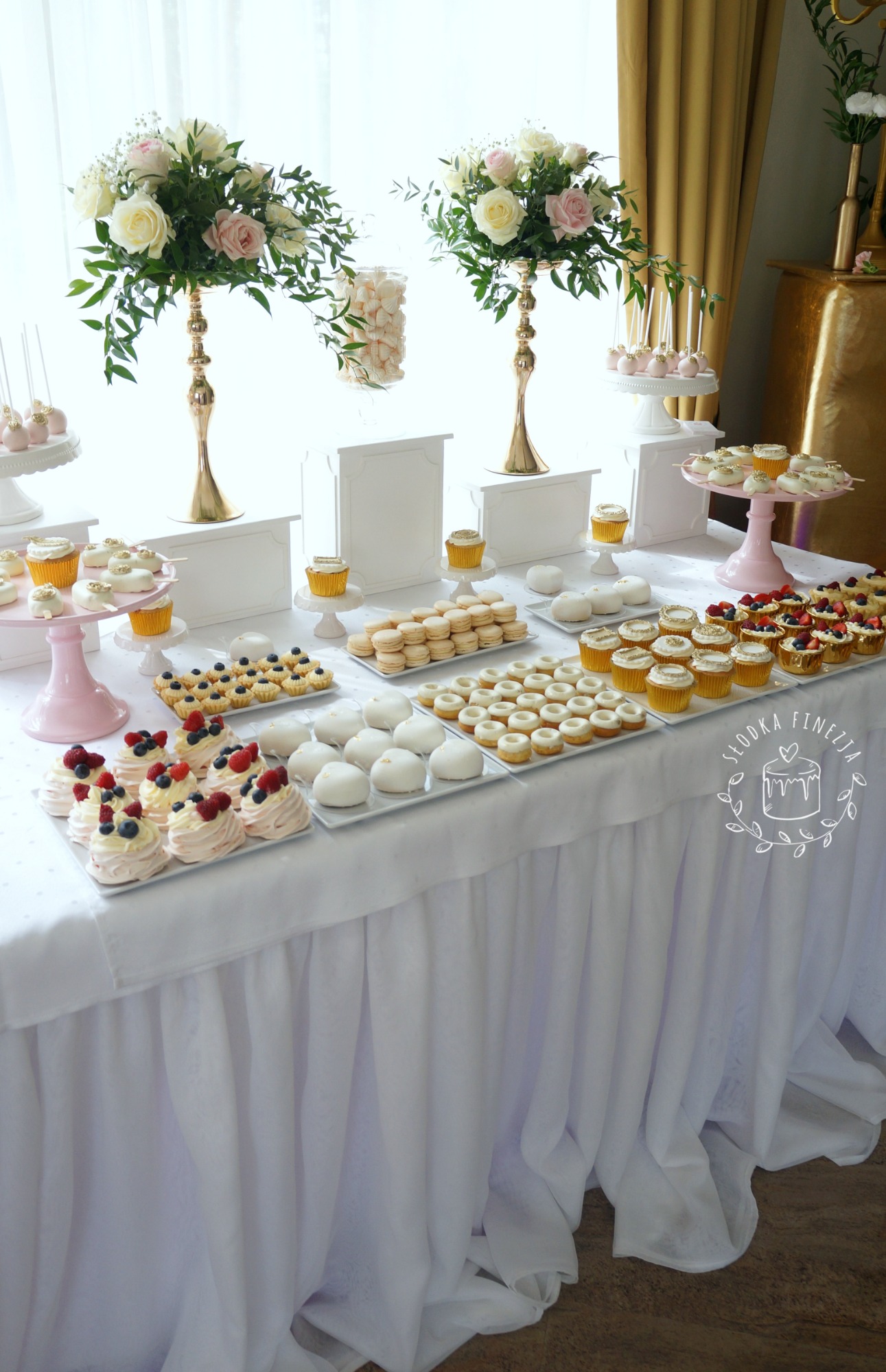 Stół z białym obrusem udekorowany różnorodnymi deserami: babeczki, bezy z owocami, makaroniki, cake pops, ustawione pod dekoracjami z kwiatów w złotych wazonach.