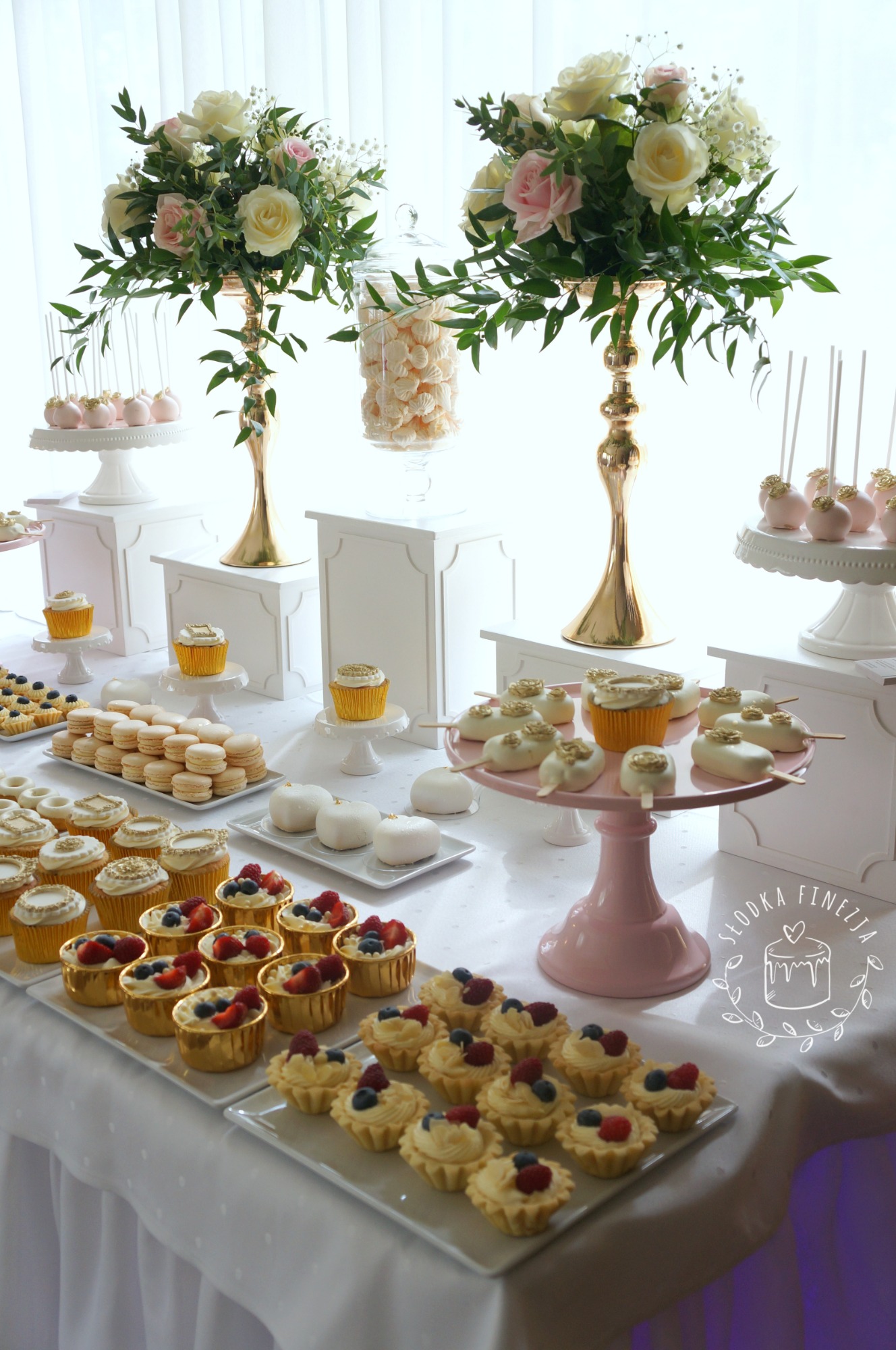 Elegancki stół z deserami na przyjęciu: babeczki z owocami, makaroniki, cake pops, beziki w szklanym słoju oraz kompozycje kwiatowe w złotych wazonach.