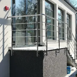 Błyszcząca, metalowa balustrada z poziomymi prętami zabezpieczająca wejście do budynku z przeszklonymi drzwiami i oknami, widoczne mocowania do schodów i ściany, cień rzucany przez pręty na elewacji.