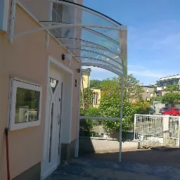 Zadaszenie nad wejściem do domu z przezroczystego poliwęglanu na białej, metalowej konstrukcji, widok z boku na tle błękitnego nieba.