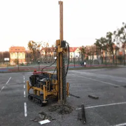 Żółty wiertniczy pojazd gąsienicowy na parkingu z widocznymi narzędziami i dokumentami, w tle budynki mieszkalne i boisko.