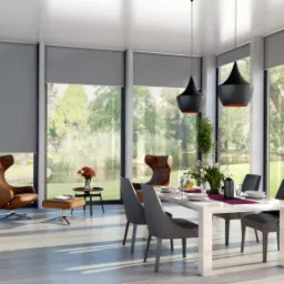 Nowoczesne wnętrze jadalni z dużymi oknami, zasłoniętymi szarymi materiałowymi osłonami przeciwsłonecznymi, widok na park z jeziorkiem.