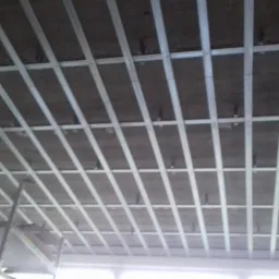 Metalowa konstrukcja podwieszanego sufitu zamocowana do surowego betonowego stropu, widok z dołu na ruszt z profili.