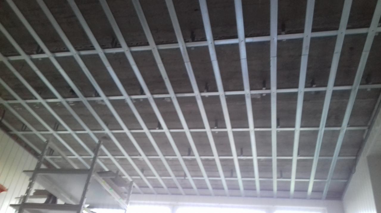 Metalowa konstrukcja podwieszanego sufitu zamocowana do surowego betonowego stropu, widok z dołu na ruszt z profili.