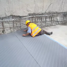 Pracownik w żółtym kasku i pomarańczowej kamizelce układa szare, perforowane maty izolacyjne na fundamencie, widoczne zbrojenie ściany fundamentowej.