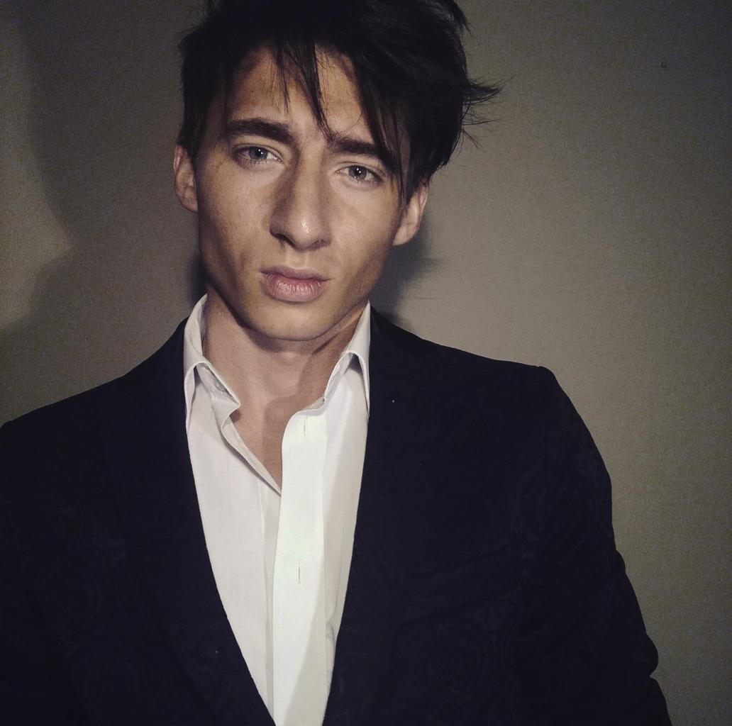 Młody mężczyzna w czarnej marynarce i białej koszuli, z ciemnymi włosami, pozuje na szarym tle. Ujęcie od przodu, oświetlenie z lewej strony.