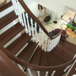 Drewniane schody spiralne z białymi balustradami, widok z góry. Pracownik w trakcie montażu, widoczny kompresor i narzędzia. Ciemne drewno, jasne tło.