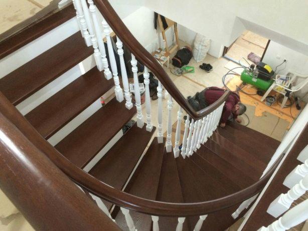 Drewniane schody spiralne z białymi balustradami, widok z góry. Pracownik w trakcie montażu, widoczny kompresor i narzędzia. Ciemne drewno, jasne tło.