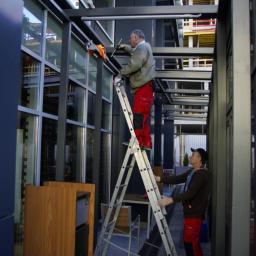 Siekielewscy - Dwóch mężczyzn myje okna na wysokości przy użyciu drabiny aluminiowej w przestrzeni między budynkami o nowoczesnej architekturze.