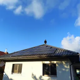 Montaż paneli słonecznych na dachu domu jednorodzinnego z szarej dachówki; pracownik w czarnej kurtce i czerwonych rękawicach na tle błękitnego nieba z chmurami.