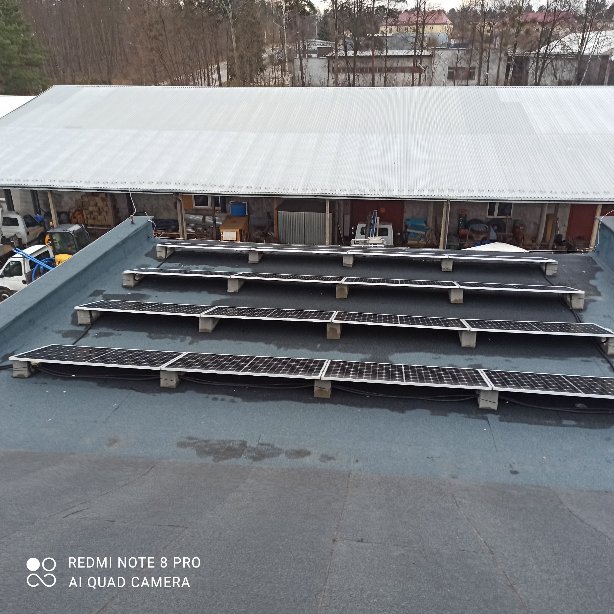 Rząd paneli słonecznych, zamontowanych na betonowych podstawach, na płaskim, pokrytym papą dachu dużego budynku gospodarczego. W tle widoczne inne budynki i drzewa.