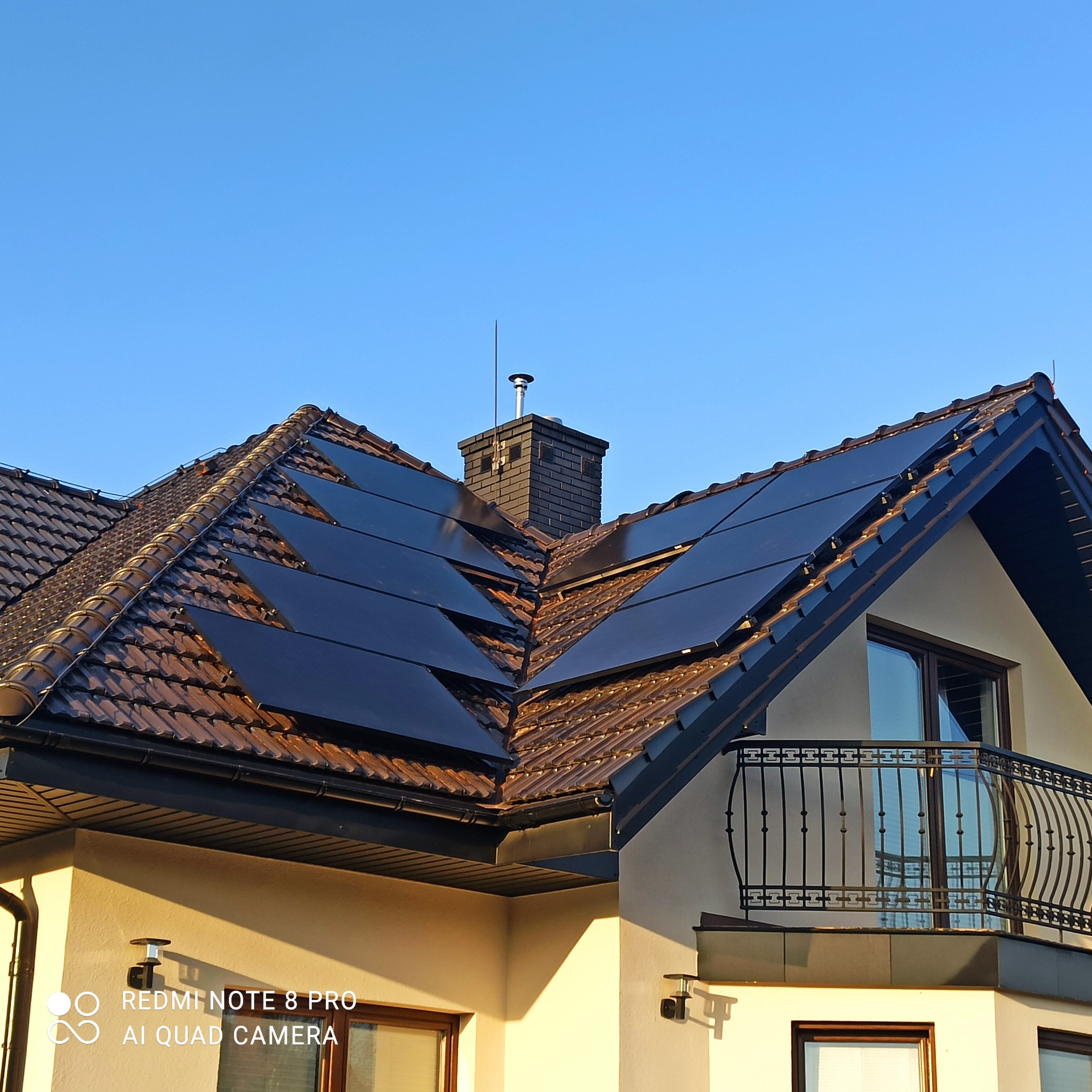 Brązowa dachówka z panelami fotowoltaicznymi na dachu domu jednorodzinnego z balkonem i ceglanym kominem na tle błękitnego nieba.
