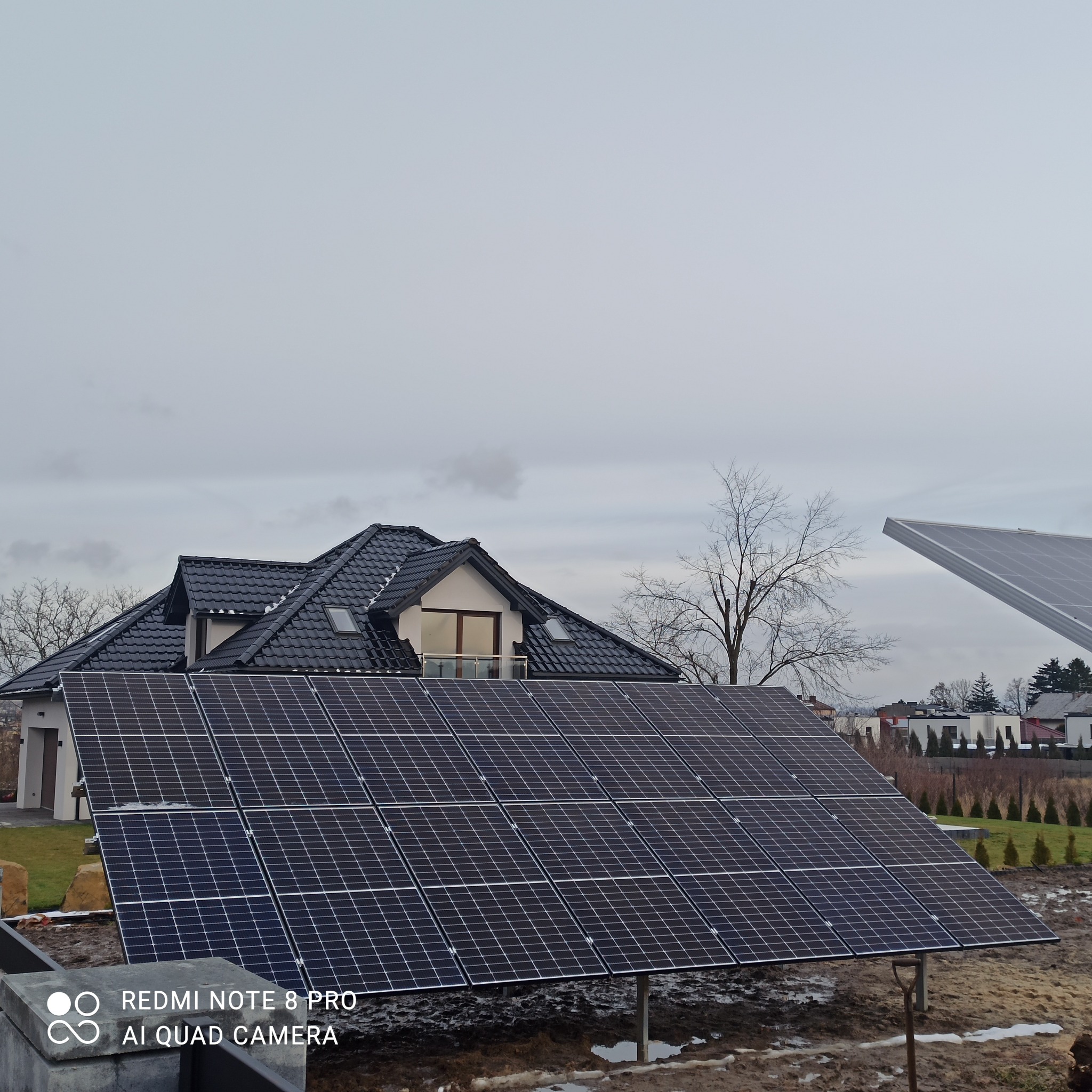 Panel słoneczny na tle domu jednorodzinnego z czarną dachówką, pochmurne niebo w tle.