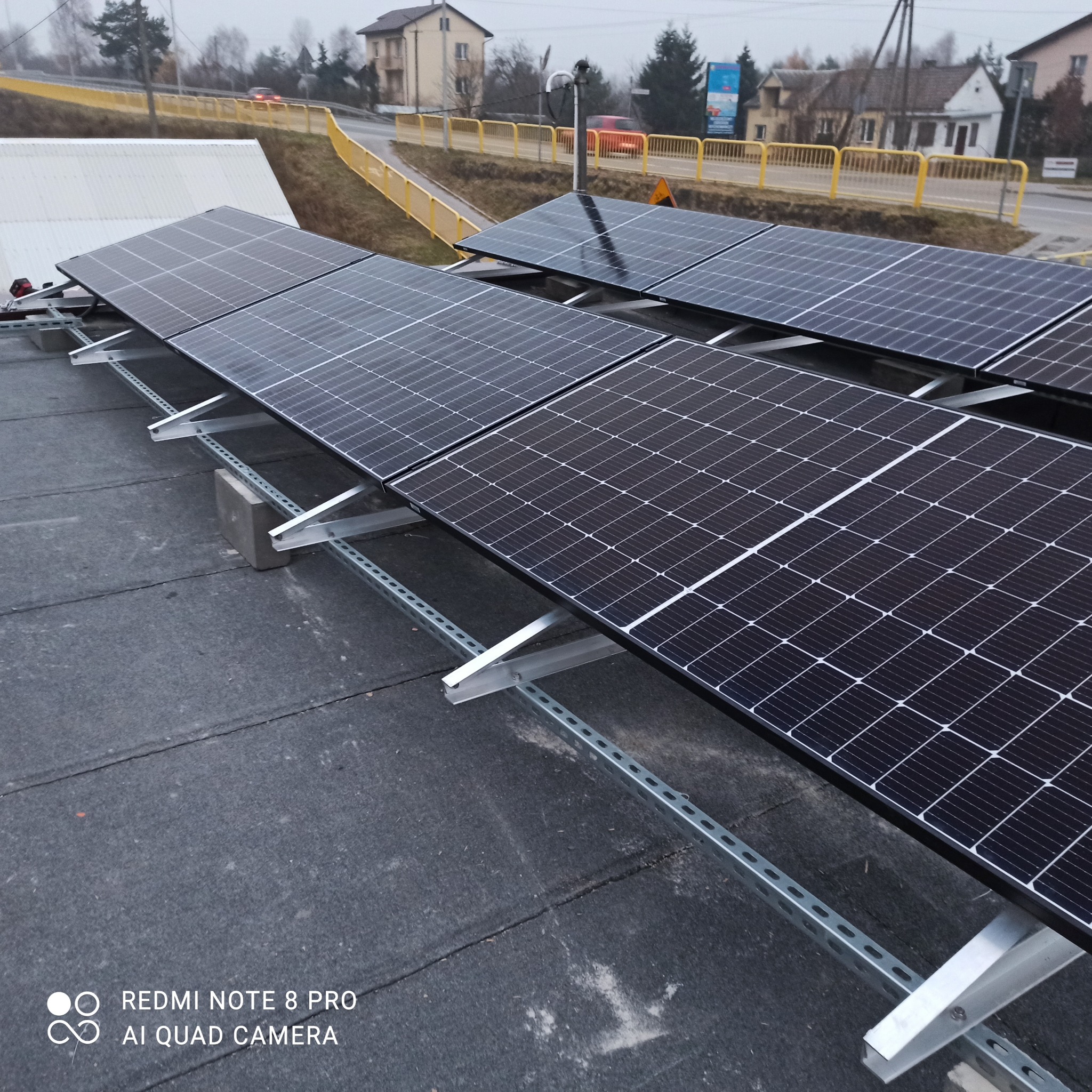 Panele słoneczne zamontowane na płaskim dachu z widoczną konstrukcją wsporczą i betonowymi obciążnikami, tło stanowią budynki i droga z barierkami.
