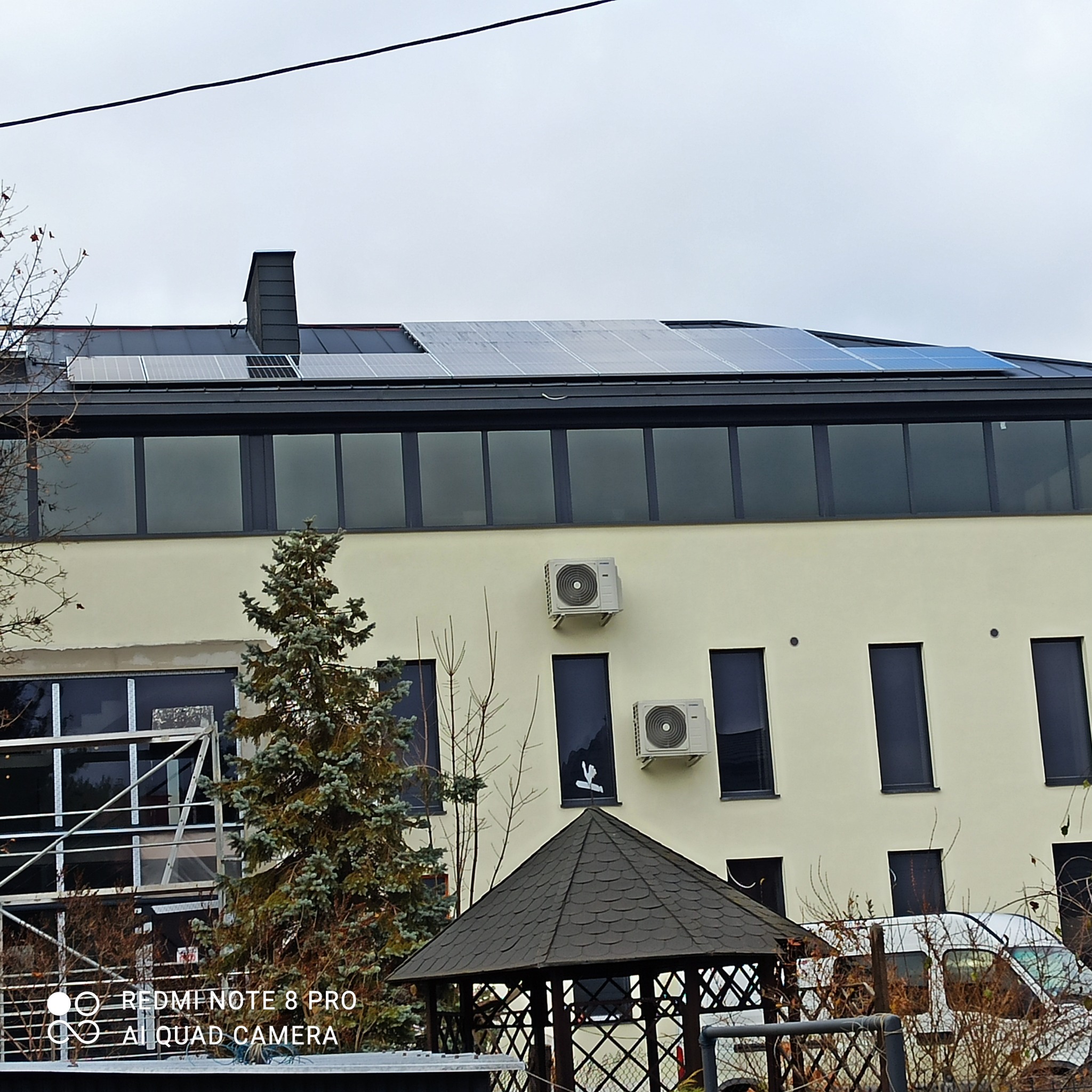 Budynek z zamontowanymi panelami fotowoltaicznymi na dachu, widoczne również klimatyzatory na elewacji i altana na pierwszym planie.