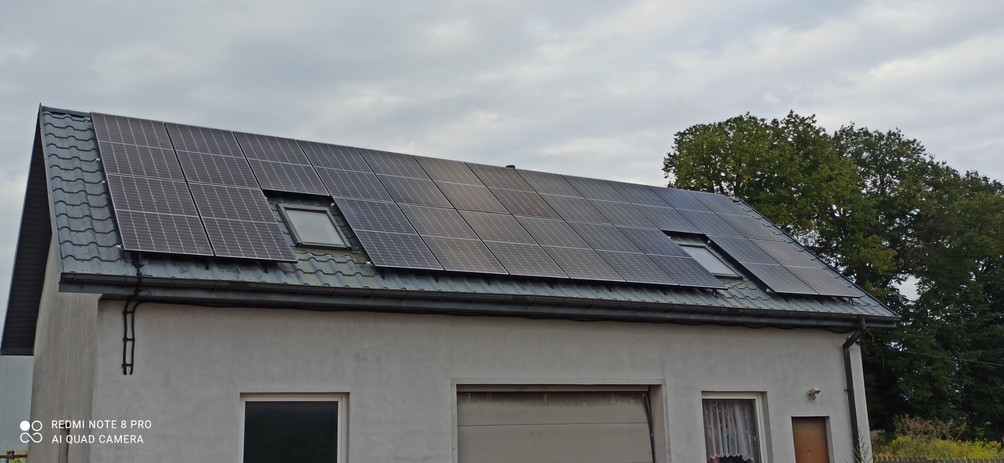 Panele słoneczne na dachu domu jednorodzinnego z dwoma oknami dachowymi, widoczne orynnowanie i fragment ogrodu w tle.