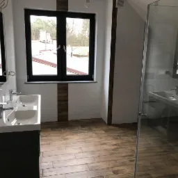 Nowoczesna łazienka na poddaszu z drewnopodobną podłogą, białymi płytkami z dekoracyjnymi wstawkami, dwiema umywalkami nablatowymi i kabiną prysznicową ze szklanymi drzwiami.