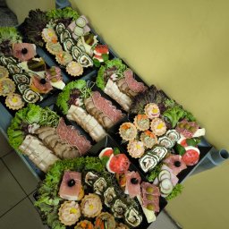 Best Food Catering - Eleganckie, czarne tace z przekąskami cateringowymi: roladki, tartaletki, wędliny, sery i warzywa, udekorowane sałatą, na metalowym wózku. Żółte tło.