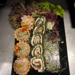 Ekskluzywne przekąski na czarnym talerzu: tartaletki z pastami i roladki z łososiem, udekorowane świeżymi sałatami i kiełkami. Elegancki catering.