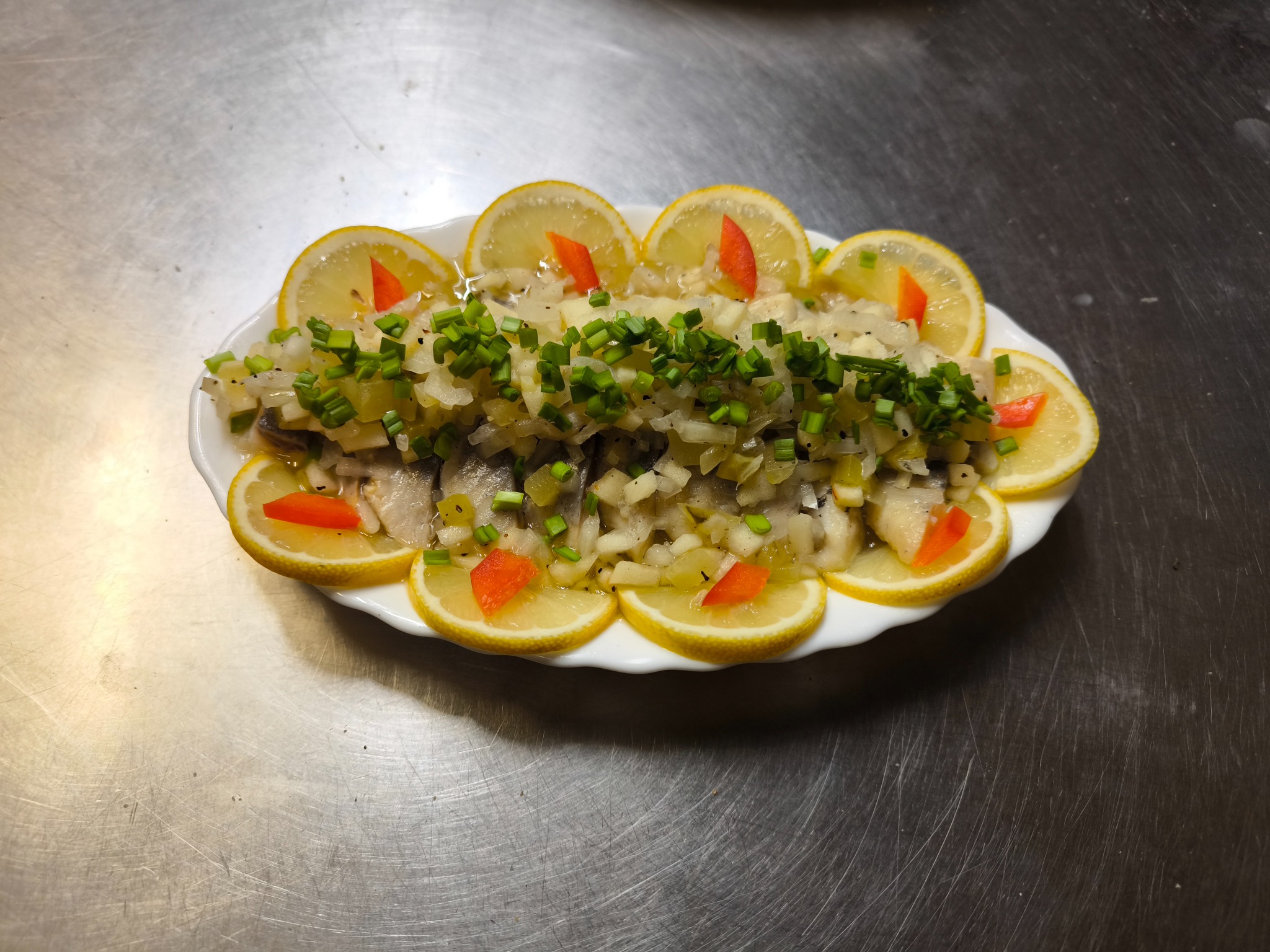 Śledź w oleju, udekorowany cytryną, cebulą i papryką, podany na białym talerzu. Widoczny na metalowym blacie.