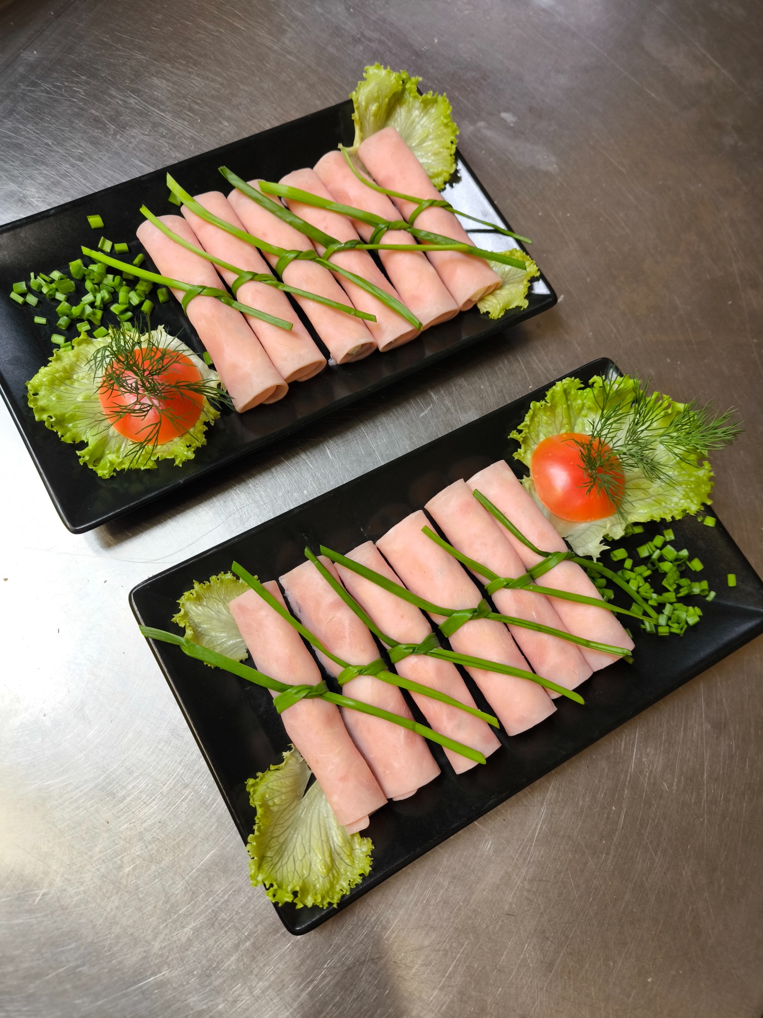 Dwa czarne talerze z roladkami z szynki, udekorowane sałatą, pomidorem i szczypiorkiem na metalowym blacie. Elegancka kompozycja przekąsek.