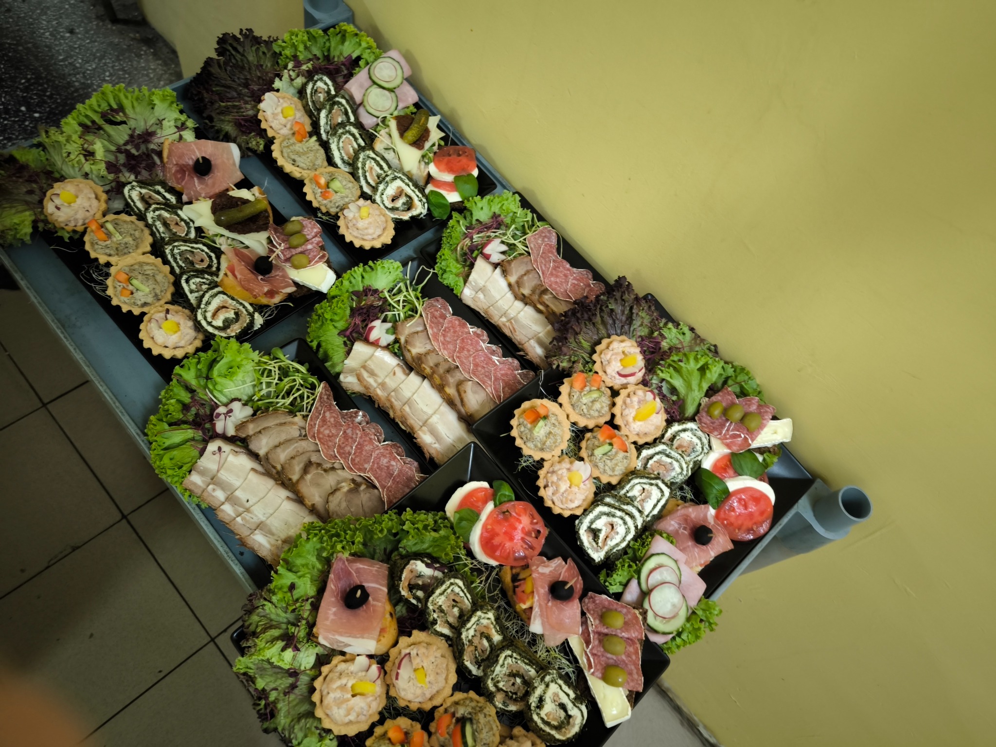 Eleganckie, czarne tace z przekąskami cateringowymi: roladki, tartaletki, wędliny, sery i warzywa, udekorowane sałatą, na metalowym wózku. Żółte tło.