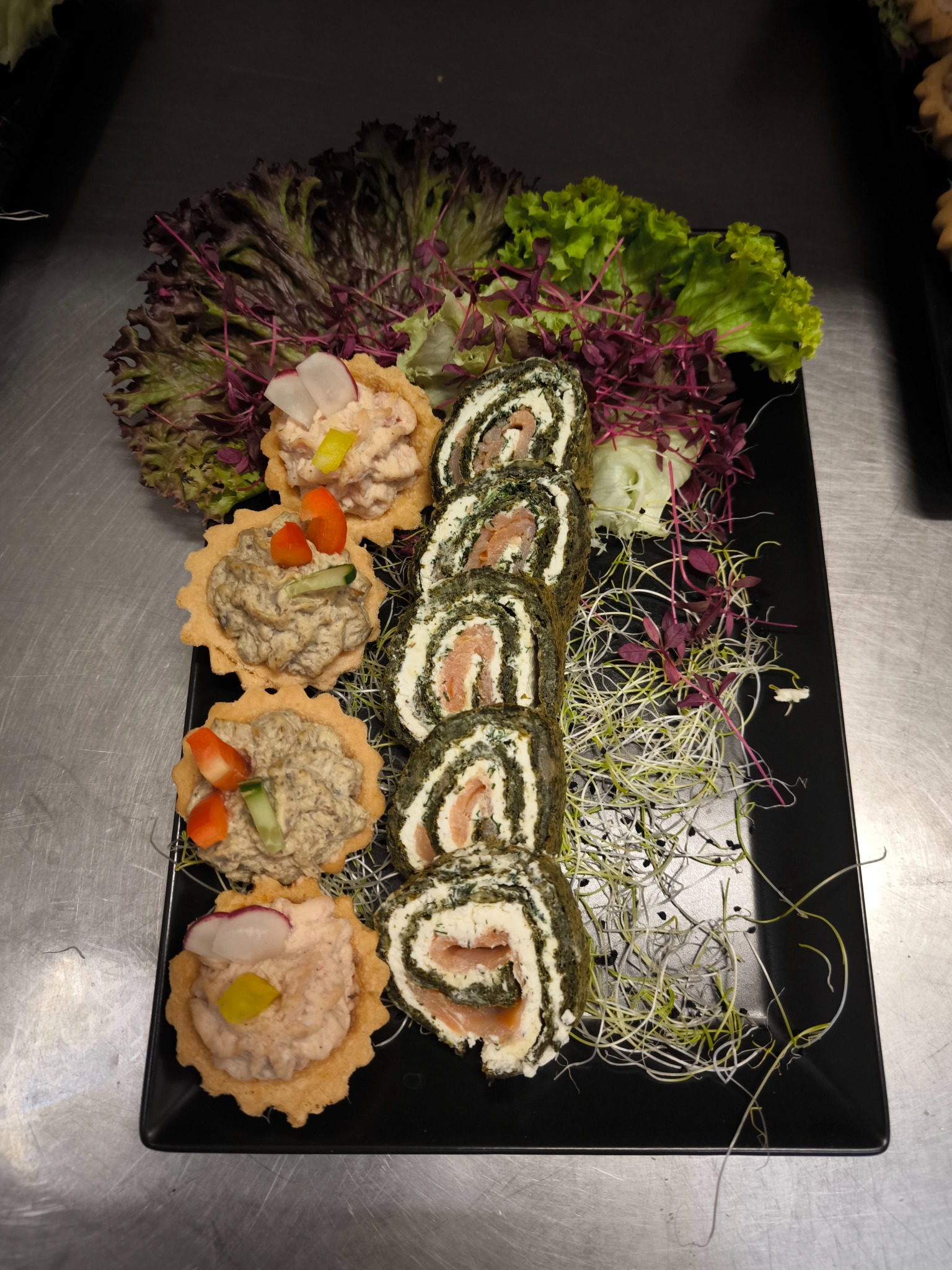 Ekskluzywne przekąski na czarnym talerzu: tartaletki z pastami i roladki z łososiem, udekorowane świeżymi sałatami i kiełkami. Elegancki catering.