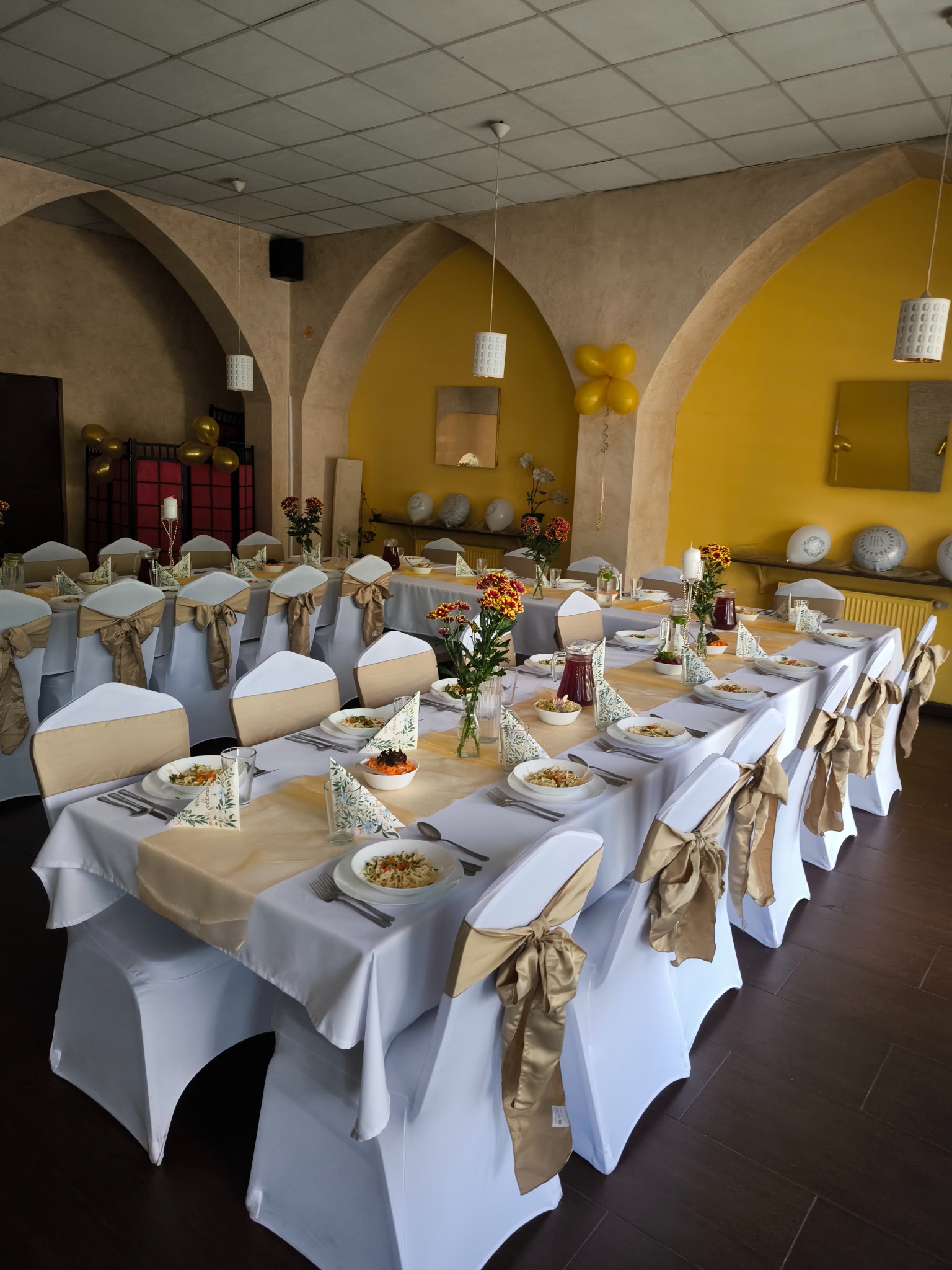 Uroczysty stół z białymi obrusami i złotymi akcentami, nakryty zastawą i dekoracjami kwiatowymi. Krzesła z białymi pokrowcami i złotymi kokardami. Żółte ściany i balony w tle.