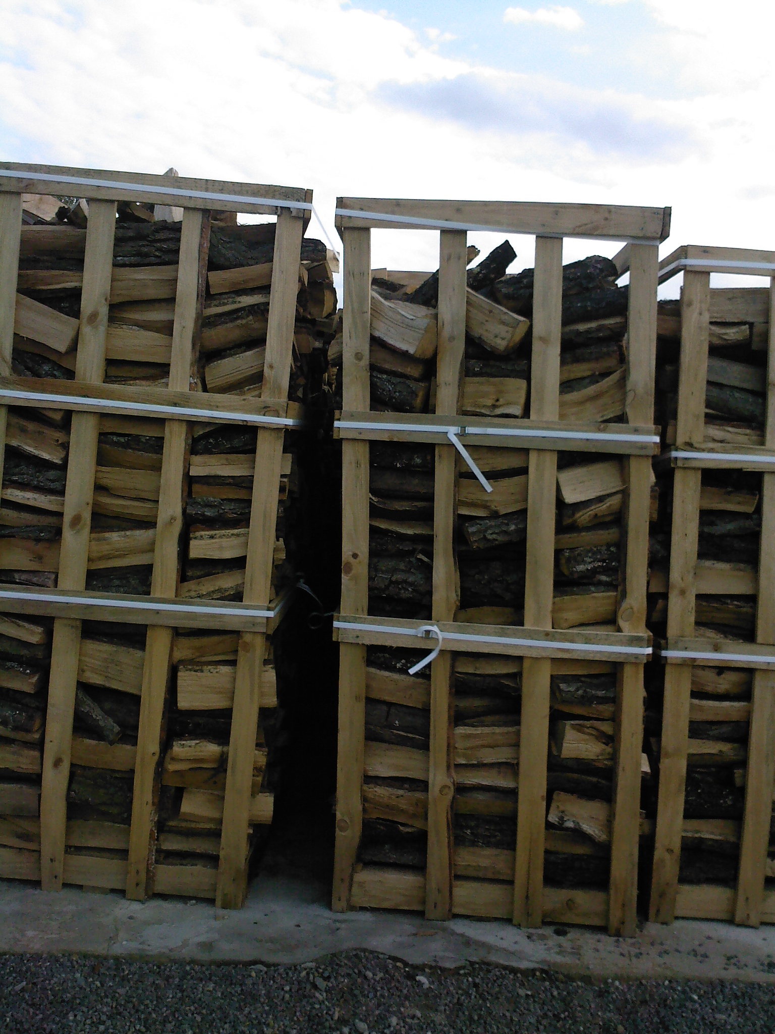 Ułożone w paletach drewno kominkowe, zabezpieczone białymi taśmami, widoczne szczeliny między paletami.