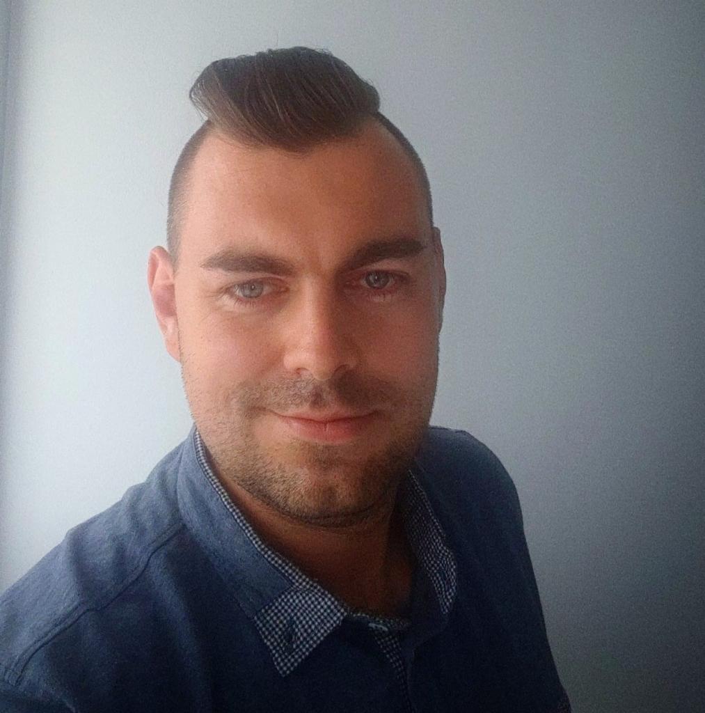 Portret mężczyzny z krótką fryzurą i lekkim zarostem, ubranego w niebieską koszulę, na tle jasnej ściany. Uśmiech na twarzy, bezpośrednie spojrzenie w obiektyw.