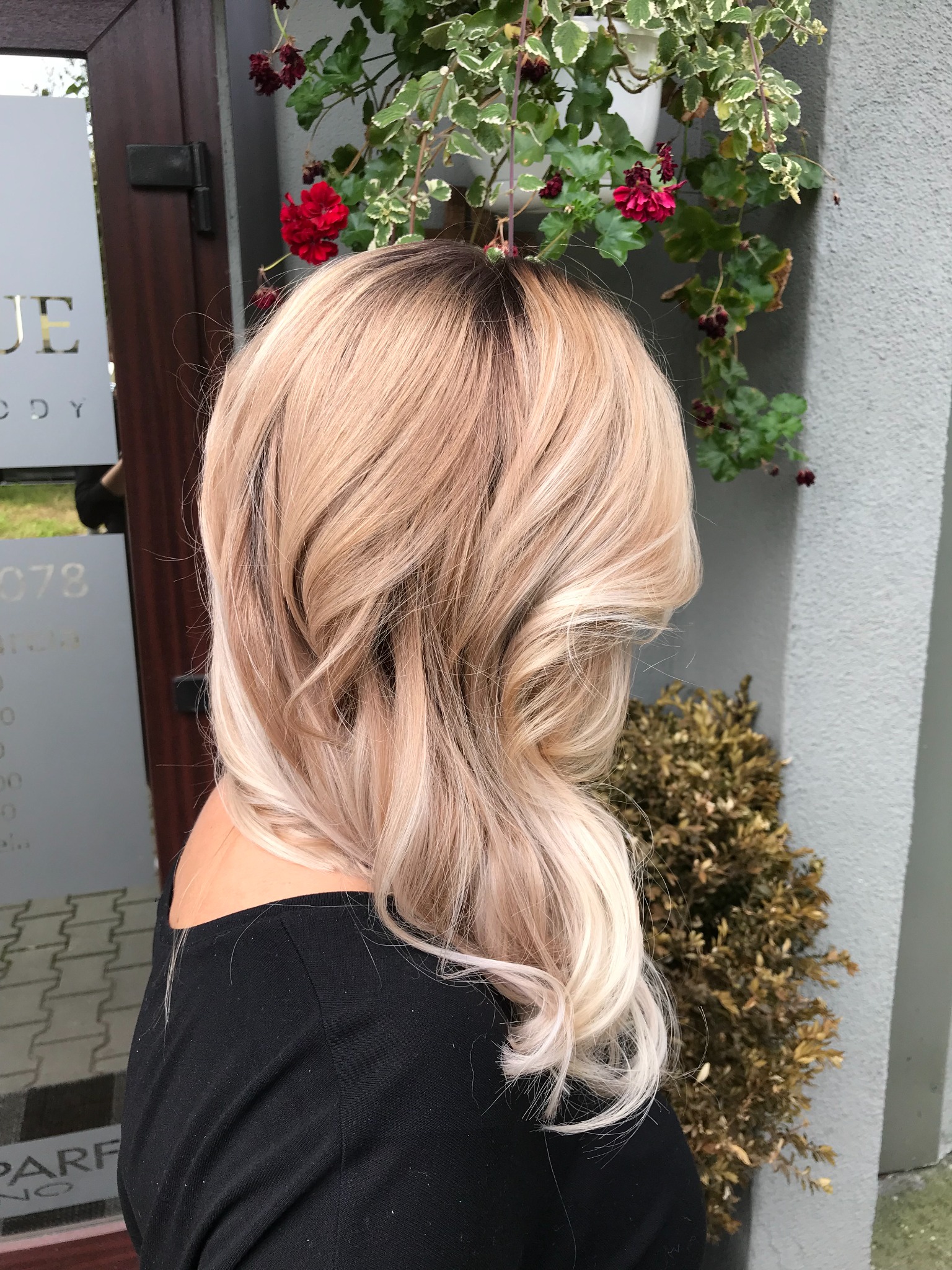 Damska fryzura w odcieniach blondu z ciemnymi odrostami, widoczna z boku na tle wejścia do salonu z dekoracją roślinną.