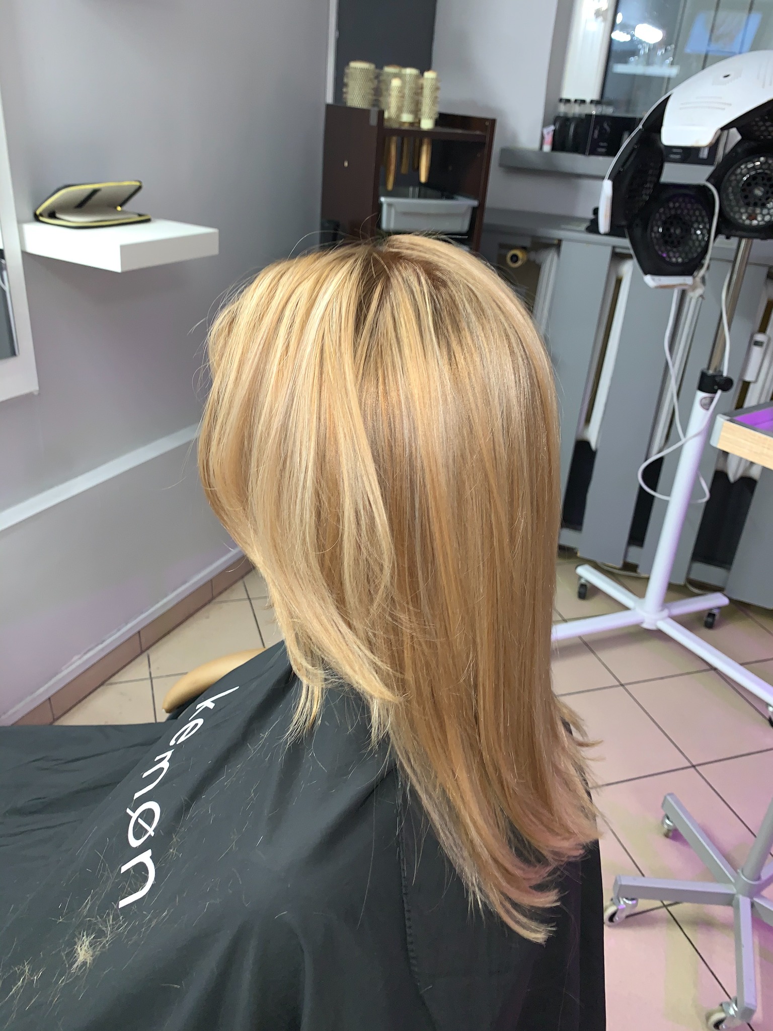 Kobieta z odwróconą głową, prezentująca świeżo ostrzyżone, blond włosy z refleksami w salonie, widoczny fartuch fryzjerski z napisem i obcięte włosy na nim.