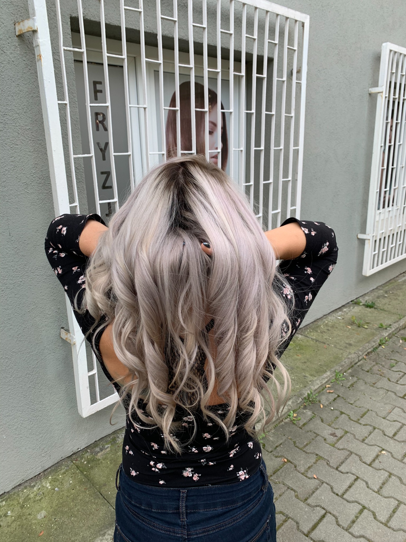 Kobieta tyłem prezentuje platynowe, pofalowane włosy z ciemniejszym odrostem, stojąc na zewnątrz budynku z napisem na szybie i kratami w oknach.