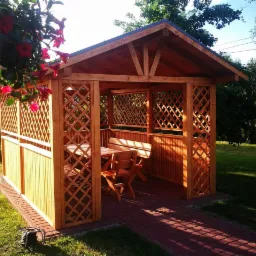 Drewniana altana ogrodowa z kratownicowymi ścianami, wyposażona w stół i ławki, otoczona zielonym trawnikiem i ścieżką z czerwonej kostki brukowej, z wiszącymi kwiatami w donicy w lewym górnym rogu.